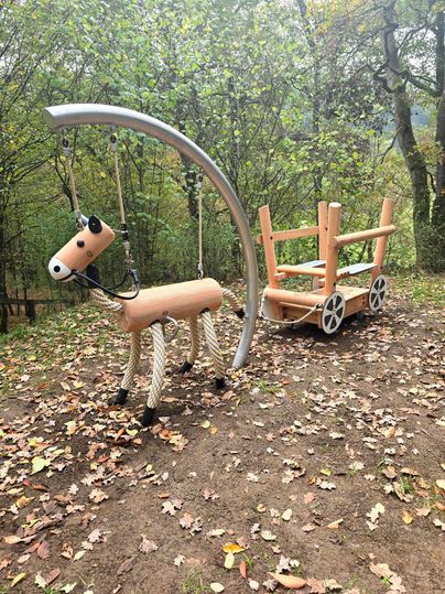 Ein Spielplatz mit einem Holzpferd und einer Kutsche. Die Umgebung ist von Bäumen und herbstlichem Laub geprägt.