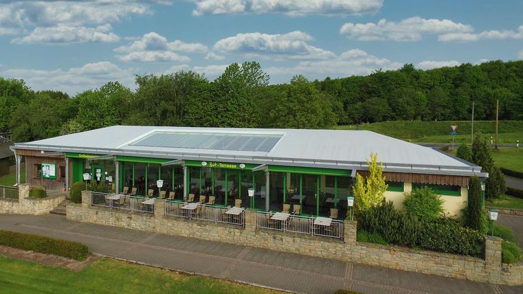 Un bâtiment moderne avec de grandes fenêtres et un toit lumineux. Il est situé au milieu d'un paysage verdoyant et dispose d'une terrasse avec des sièges en plein air.