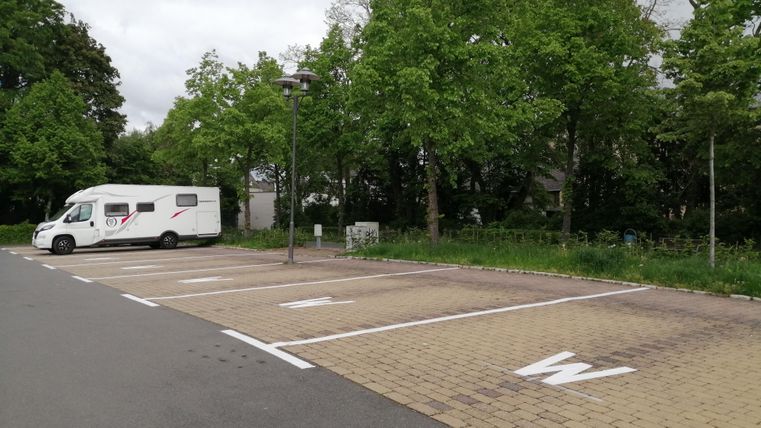 Een camper op een parkeerplaats met veel vrije staanplaatsen. De omgeving is groen met bomen.
