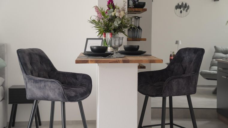 A modern dining area with two comfortable gray stools and a rustic table. On the table are plates and a vase of fresh flowers.