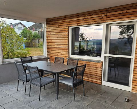 Ein gemütlicher Außenbereich mit einem Tisch und Stühlen auf einer Terrasse. Man sieht große Fenster und eine grüne Umgebung.