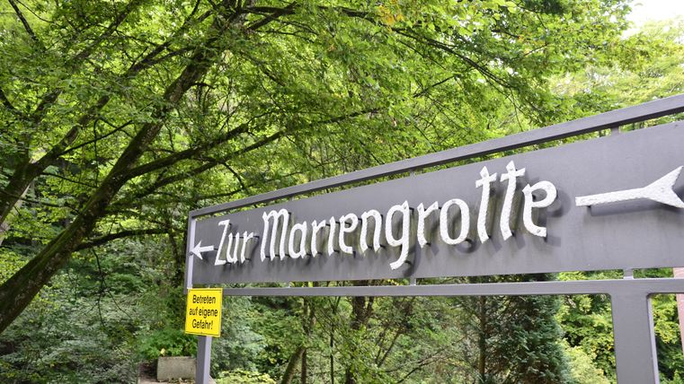 Ein Wegweiser zur Mariengrotte in einer grünen Umgebung. Umgeben von Bäumen und natürlicher Vegetation.