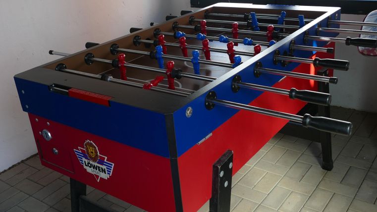 A table football game in a room with windows. The table is red and blue and stands on a tiled floor.