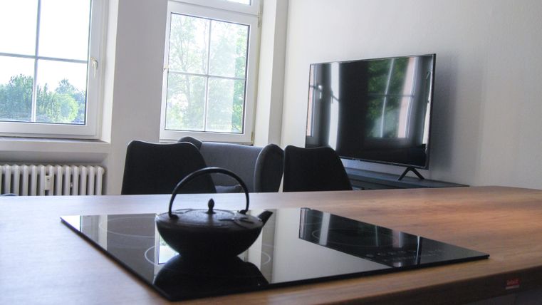 Eine moderne Küche mit einem Holz-Esstisch und einem Kochfeld. Im Hintergrund sind ein Fernseher und Fenster, die viel Licht hereinlassen.