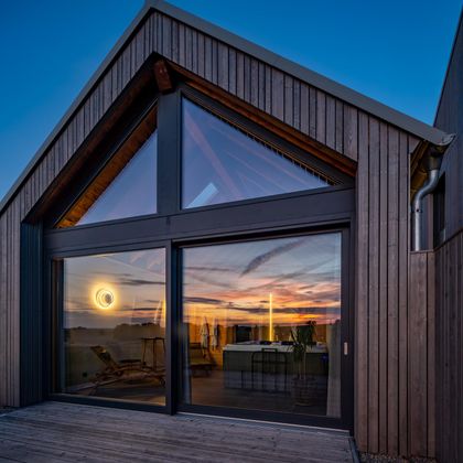 Ein modernes Holzhaus mit großen Fenstern und einem Blick auf den Sonnenuntergang. Der Innenraum ist hell und gemütlich, mit sanften Beleuchtungseffekten.