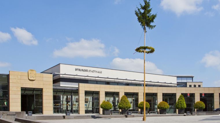 Ein modernes Gebäude mit großen Fenstern und einer hellen Fassade. Umgeben von begrünten Bereichen und einem klaren blauen Himmel.