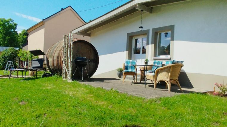 Een gezellige buitenruimte met een tafel en stoelen. Op de achtergrond staat een groot houten vat en het gras is groen.