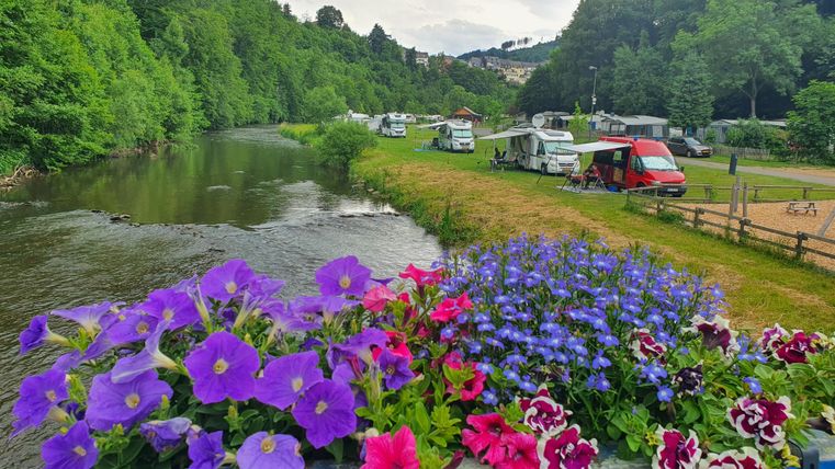 Ein malerischer Campingplatz am Fluss mit grünen Wäldern im Hintergrund. Bunte Blumen schmücken den Vordergrund und verleihen der Szene Farbe.