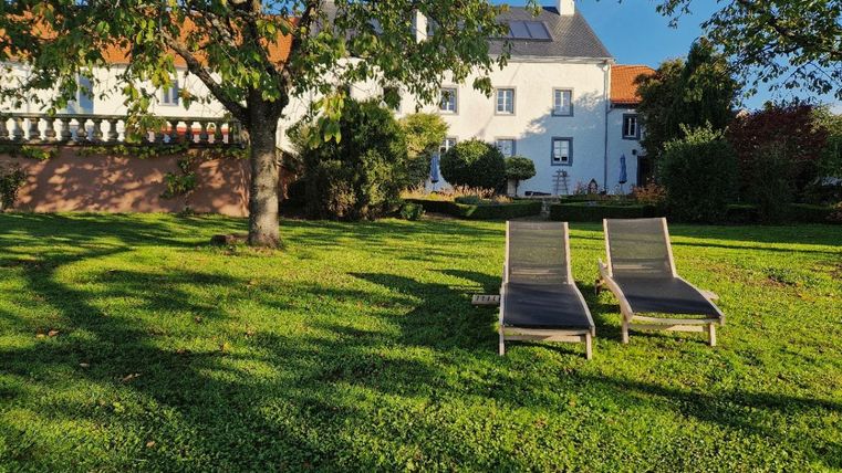 Twee ligstoelen op een groene weide voor een wit huis. Op de achtergrond zijn bomen en struiken zichtbaar.