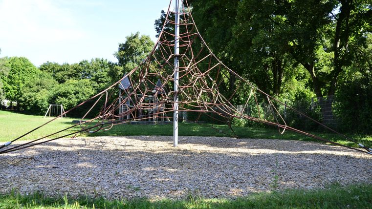 A playground with a climbing frame made of ropes is located on a meadow. In the background, trees and green grass are visible.