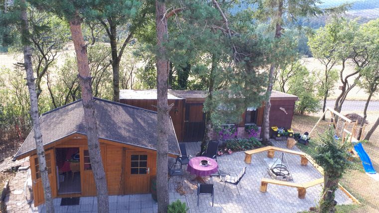 Une maison de vacances confortable entourée d'arbres. Devant se trouvent une terrasse avec des sièges et une aire de jeux.