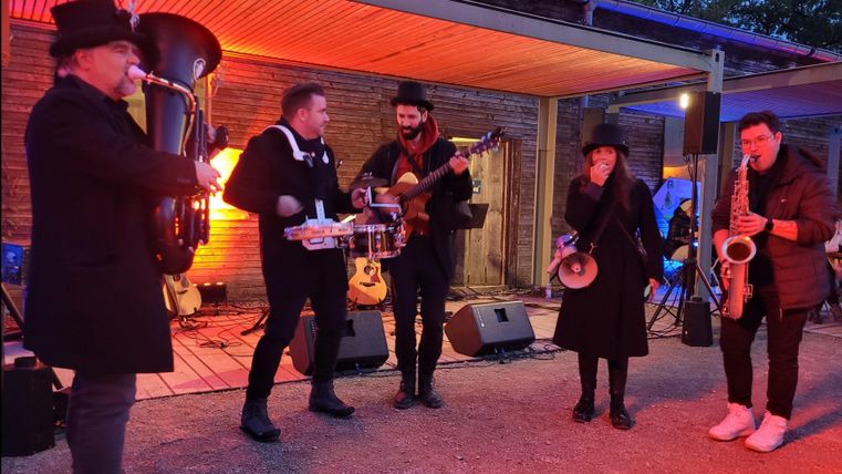 Eine Band spielt live Musik in einem gemütlichen Außenbereich. Die Künstler sind in schwarz gekleidet und es gibt farbiges Licht im Hintergrund.