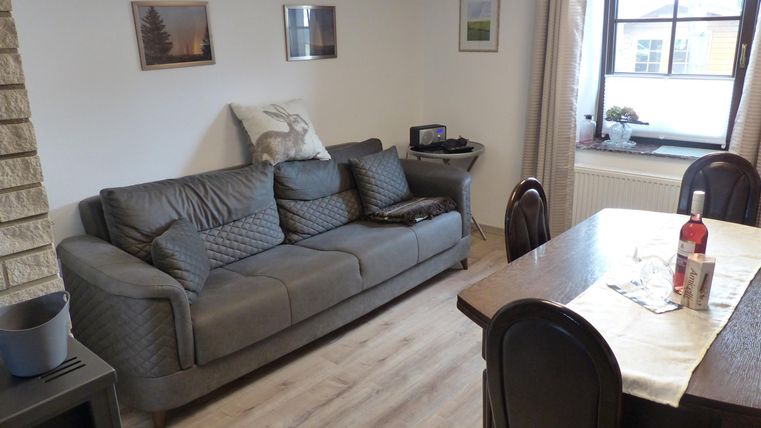 Un salon confortable avec un canapé gris et une table à manger. Les murs sont décorés de tableaux, et une fenêtre laisse entrer la lumière du jour.