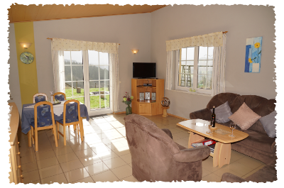Un salon confortable avec une table, des chaises et un canapé. De grandes fenêtres laissent entrer beaucoup de lumière et offrent une vue sur les environs.