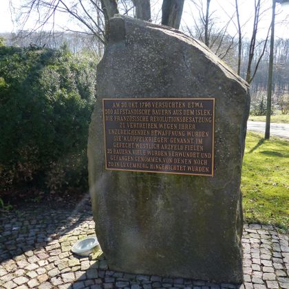 Ein großer Steinschinz mit einer Inschrift in deutscher Sprache. Der Stein erinnert an ein historisches Ereignis und befindet sich in einer grünen Umgebung.