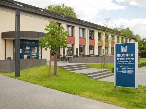 A modern library building with an inviting entrance. There are trees on the premises and an information board with opening hours.