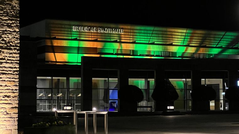 A modern building at night, illuminated with green and orange lights. The façade is simple yet appealingly designed.