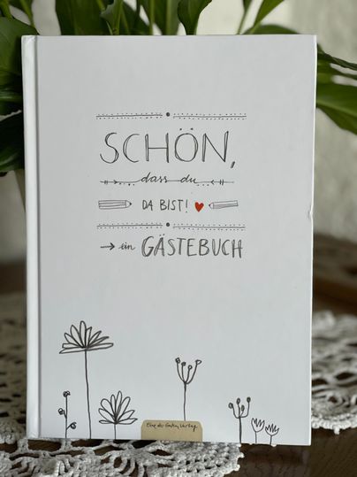 Een uitnodigend gastenboek met de tekst "LEUK dat je er bent!" en een eenvoudige tekening van bloemen. Het ligt op een tafel, omringd door een decoratieve haakwerk.