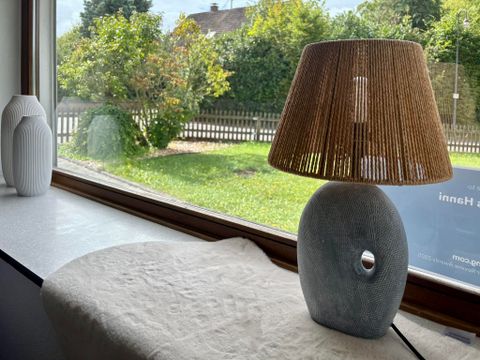 Eine stilvolle Lampe mit einem runden, grauen Sockel und einem schilfgefertigten Lampenschirm steht auf einem Tisch. Draußen ist eine grüne Wiese und ein Zaun zu sehen.