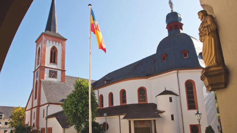 Eine malerische Kirche mit einem hohen Turm und buntem Dach. Im Vordergrund steht eine Fahne und es sind grüne Bäume zu sehen.