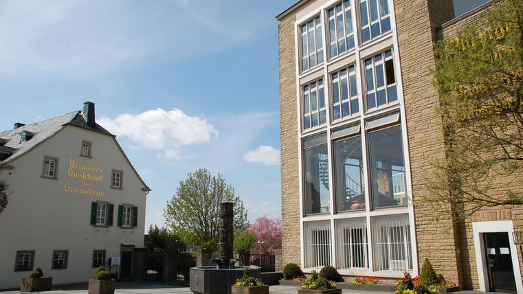 Ein heller Platz mit modernen und traditionellen Gebäuden. Bäume und Blumenkübel verleihen dem Ort eine einladende Atmosphäre.