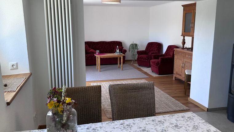 Ein gemütliches Wohnzimmer mit roten Sofas und einem Holztisch. Im Vordergrund steht ein Tisch mit einer Vase und frischen Blumen.