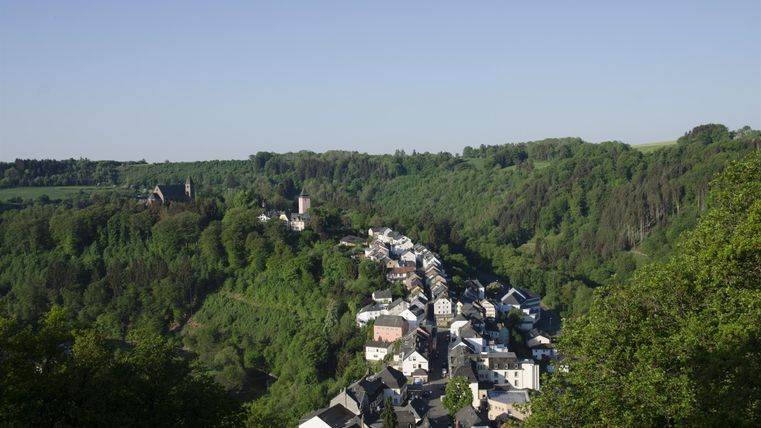 Eine malerische Landschaft mit einem kleinen Dorf, umgeben von üppigem Grün. Im Hintergrund sind Hügel und Bäume sichtbar, die dem Bild eine friedliche Atmosphäre verleihen.