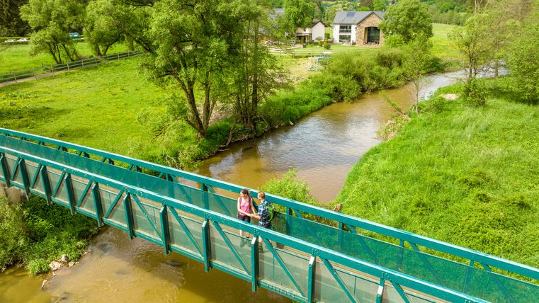 Zwei Personen auf einer grünen Brücke über einen Fluss in einer ländlichen Landschaft.