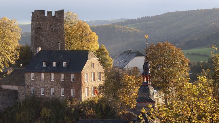 Eine historische Gebäudeansicht mit einem alten Turm und bunten Herbstbäumen. Im Hintergrund erstreckt sich eine sanfte Hügellandschaft.