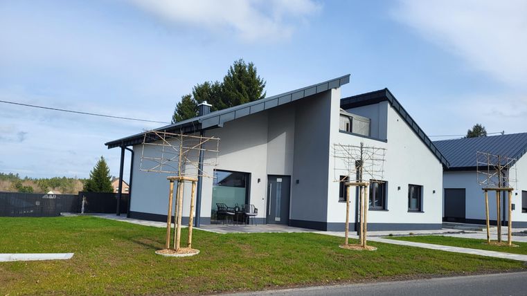 Une maison individuelle moderne avec une façade lumineuse et un toit en pente. Le jardin est bien entretenu et il y a une terrasse extérieure.