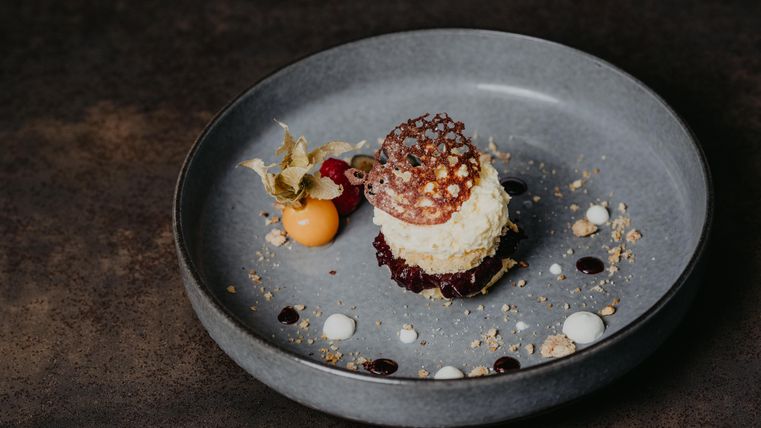 Un dessert artistiquement disposé sur une assiette grise. Il se compose d'une crème, de fruits et de sauces décoratives.