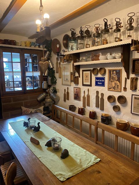 Une pièce chaleureuse avec une grande table en bois et une décoration nostalgique sur les murs. De vieilles lampes et des ustensiles de cuisine sont disposés de manière décorative.