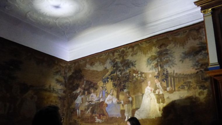 Ein historischer Raum mit einer dekorativen Wandmalerei. Personen stehen im Vordergrund und betrachten die kunstvolle Darstellung.