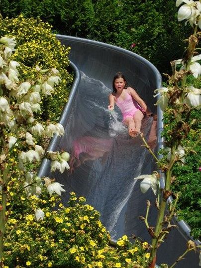 Ein Mädchen rutscht fröhlich eine Wasserrutsche hinunter. Umgeben von bunten Blumen und Bäumen.
