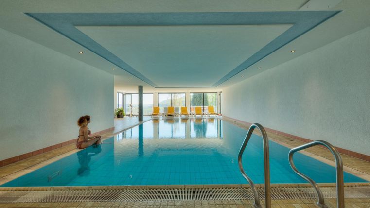Ein ruhiges Schwimmbad mit klar blauem Wasser und großen Fenstern. Eine Person sitzt am Beckenrand und genießt die entspannende Atmosphäre.