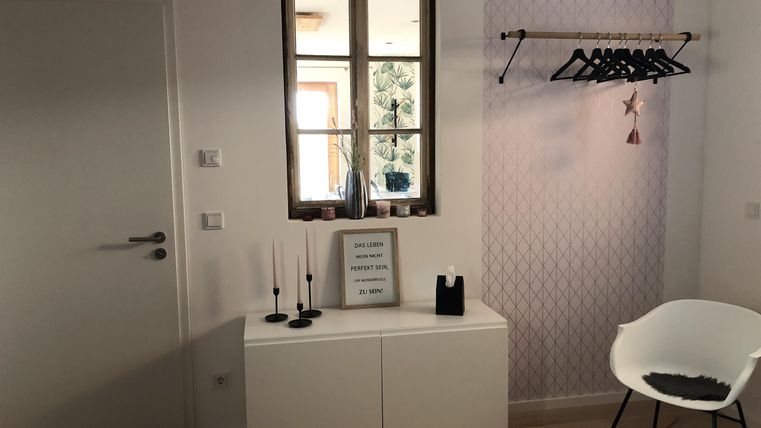 Ein heller Raum mit einem Fenster und einem weißen Sideboard. An der Wand hängt eine Kleiderstange mit Kleidungsstücken.