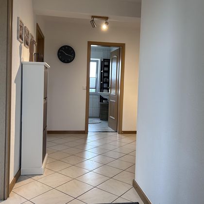 Een eenvoudige gang met een tegelvloer. Aan de linkerkant staat een witte kast en op de achtergrond zie je een open deur naar een kamer.