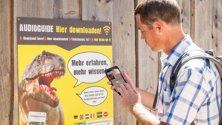 Een man scant met zijn smartphone een QR-code op een poster. De poster bevat informatie over audiogidsen en toont een afbeelding van een dinosaurier.