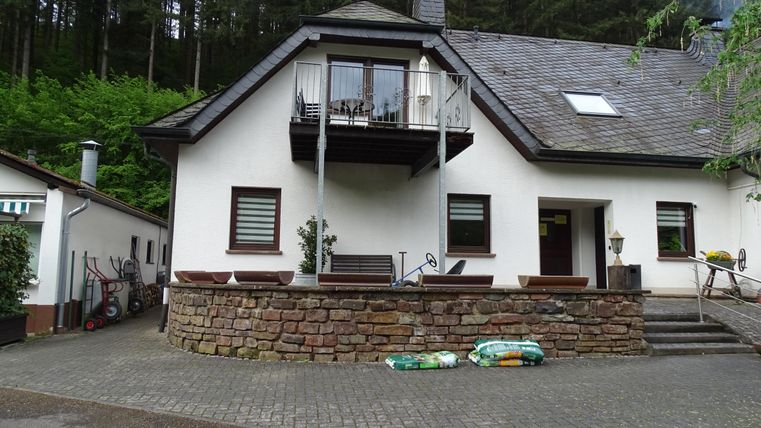 Une maison à deux étages dans un environnement rural avec un balcon. À l'arrière-plan, on voit des arbres, et l'extérieur est bien entretenu.