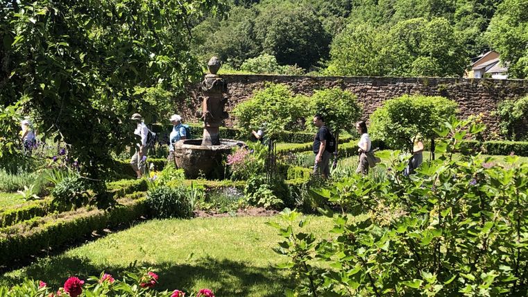 Ein schöner Garten mit einer kleinen Quelle und bunten Blumen. Menschen genießen die Natur vor einer malerischen Hintergrundlandschaft.