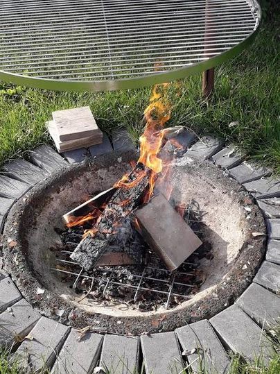 Ein romantisches Lagerfeuer in einer Feuerstelle mit einem Grillrost darüber. Rundherum wächst frisches Gras und einige Holzstücke liegen bereit.