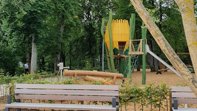 Un terrain de jeu dans un parc avec des arbres verts. À l'arrière-plan se trouve une structure de jeu colorée et des bancs invitent à s'asseoir.