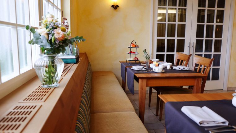 Un coin repas confortable avec des tables et des chaises en bois. Sur le rebord de la fenêtre se trouve un vase à fleurs et un petit présentoir à desserts.