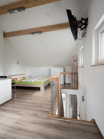 Une chambre lumineuse avec des plafonds en bois, une télévision au mur et un lit double. Il y a un escalier et un petit coin salon.