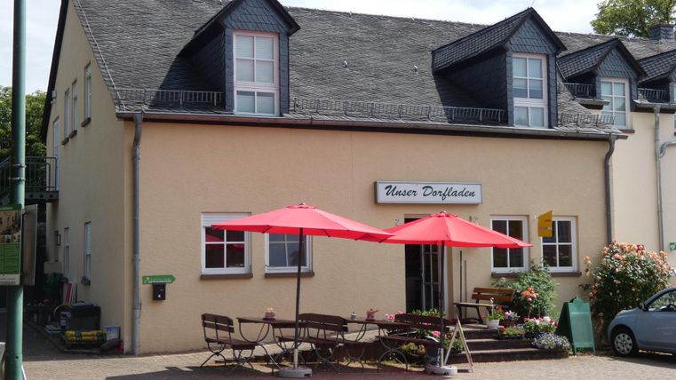 Un bâtiment confortable avec une enseigne indiquant "Maison de la Trinité". Devant la maison, il y a des tables avec des parasols rouges et des fleurs.