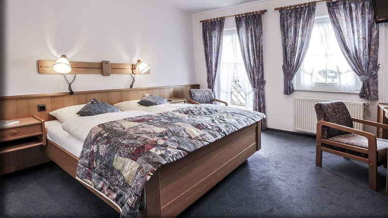 Une chambre confortable avec un grand lit et une chaise. Les fenêtres sont ornées de rideaux, et la pièce a un charme chaleureux et accueillant.