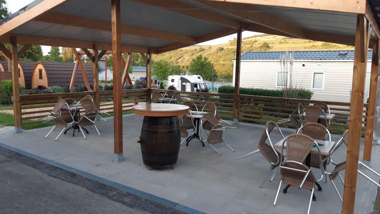 Ein überdachter Außenbereich mit Tischen und Stühlen. Im Hintergrund sind einige Campingunterkünfte zu sehen.
