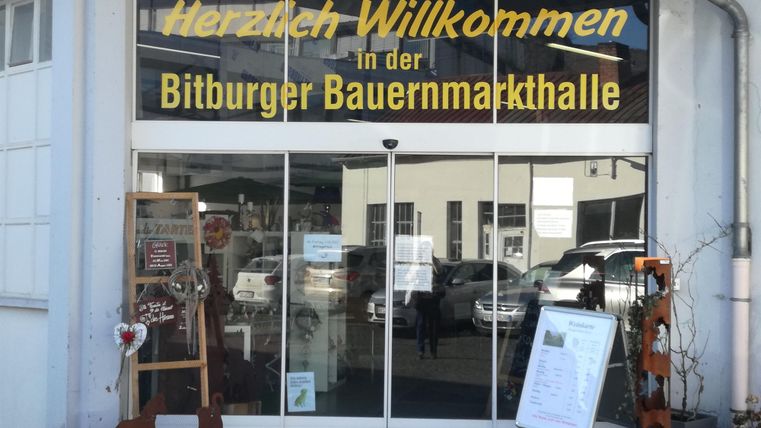 Une entrée accueillante vers la Halle du Marché Paysan de Bitburg. Les vitrines sur le côté affichent des informations et des décorations.