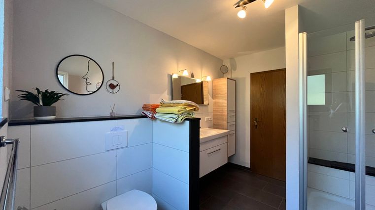 Ein modernes Badezimmer mit einem WC, einer Dusche und hellem Licht. Auf der Ablage liegen sauber gefaltete Handtücher und es gibt eine dekorative Pflanze.