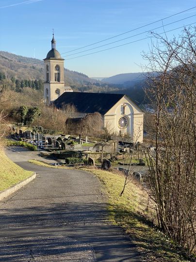 Eine ruhige Landschaft mit einer Kirche und einem Friedhof. Im Hintergrund sind sanfte Hügel und ein klarer Himmel zu sehen.